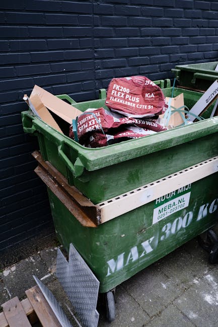 A large green wheelie bin filled with several red and black bags of construction and building materials, positioned on a concrete pavement next to a dark brick wall. The bags are labelled with product
